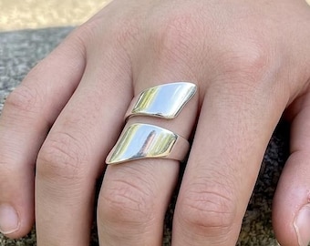 May include: A polished silver-tone ring featuring an open, bypass-style design with two smooth, tapered, and slightly curved metal segments that wrap around the finger, creating a sleek and reflective surface with a modern, minimalist aesthetic.
