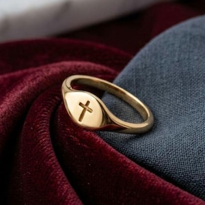 May include: A gold signet ring featuring a small, engraved cross symbol on the flat, oval-shaped face. The ring has a polished, metallic finish and a smooth, rounded band. The text "Handcrafted 18k Gold Cross Ring" is printed at the bottom.