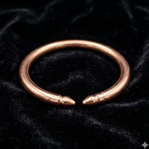 Peut inclure: Un bracelet manchette en métal de couleur cuivre avec une finition lisse et polie. Le bracelet présente un design circulaire ouvert avec deux extrémités effilées en forme de balle, créant une structure de bande arrondie continue.