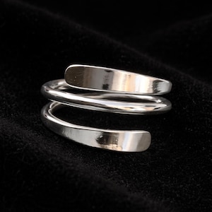 Könnte beinhalten: Ein Ring aus poliertem, silberfarbenem Metall mit einem Bypass-Design in Form einer durchgehenden Spirale. Das Band hat eine glatte, reflektierende Oberfläche, die sich um sich selbst wickelt und ein geschichtetes Aussehen mit zwei offenen Enden erzeugt.