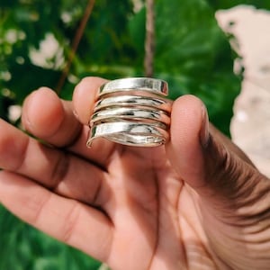 Puede incluir: Un anillo de tono plateado con un diseño de envoltura en espiral de varias capas. La banda consta de varias bobinas superpuestas que crean una apariencia continua y apilada. La superficie metálica tiene un acabado pulido y brillante.