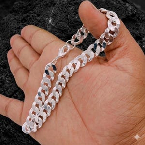 Può includere: Un bracciale a catena a maglia a catena argentata con chiusura a moschettone. Il bracciale è tenuto in una mano, mostrando le maglie lucide. La catena è un design classico, adatto all'uso quotidiano. Le maglie del bracciale sono larghe circa 0,8 cm.