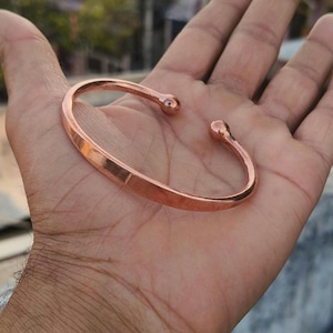 Könnte beinhalten: Ein kupferfarbenes, offenes Armband mit abgerundeten Enden. Das Armband wird in einer Hand gehalten und zeigt seine glatte, polierte Oberfläche. Das Design ist schlicht und elegant.
