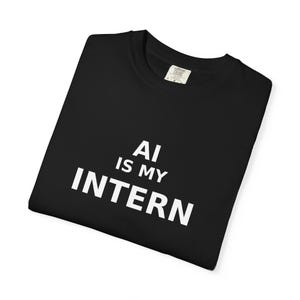 May include: Black t-shirt with the white text "AI IS MY INTERN". The shirt has a crew neck and a tag on the upper right side. The shirt is folded and appears to be made of a soft material.