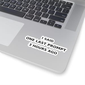 May include: A white sticker with the text "I SAID ONE LAST PROMPT 3 HOURS AGO" in black, placed on a silver laptop. The laptop keyboard is visible, with black keys and white lettering.