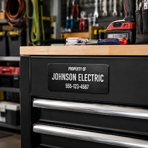 May include: A rectangular black metal identification plate attached to a black tool chest drawer. The plate features white text that reads PROPERTY OF JOHNSON ELECTRIC followed by the phone number 555-123-4567 in a clean, sans-serif font.