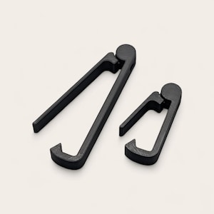 May include: Two black, textured music stand clips. One clip is larger than the other. The clips have a curved design with a circular hinge at the top and a squared-off base. The clips are designed to hold sheet music.