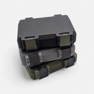 Puede incluir: Una pila de tres pequeñas cajas de almacenamiento rectangulares en tonos grises, negro y verde oliva. Cada caja tiene una superficie texturizada, bisagras negras y cierres verde oliva. Las cajas están cerradas.