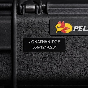 Peut inclure: Gros plan sur une valise Pelican noire avec une surface texturée. La valise comporte une plaque signalétique noire avec le nom "JONATHAN DOE" et un numéro de téléphone. Le logo Pelican est visible.
