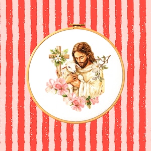 Pode incluir: Um bastidor de bordado em ponto cruz com uma imagem religiosa. A imagem retrata uma figura com cabelo comprido e barba segurando um cordeiro, rodeada por flores e uma cruz. O fundo é um padrão de listras vermelhas e brancas.
