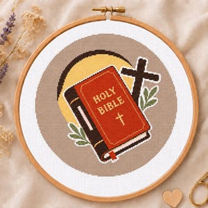May include: A cross-stitch pattern in a wooden embroidery hoop featuring a red Holy Bible with gold text, a dark brown cross, and a yellow sun circle on a beige background. The design includes small green leaf accents surrounding the central book.