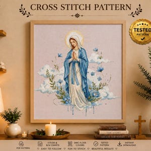 Pode incluir: Um padrão de ponto cruz emoldurado apresentando uma ilustração central de uma figura com túnica azul e branca com um halo dourado, rodeada por flores azuis e nuvens. O texto CROSS STITCH PATTERN e TESTED PATTERN aparece acima.