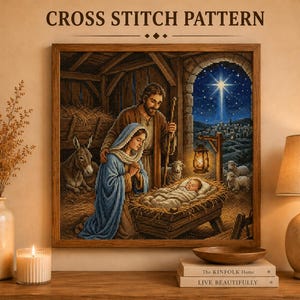 May include: A framed cross stitch pattern depicting a nativity scene with a man and woman kneeling beside a manger containing a baby. The scene includes a donkey, sheep, a glowing lantern, and a bright star in the night sky above a distant town.