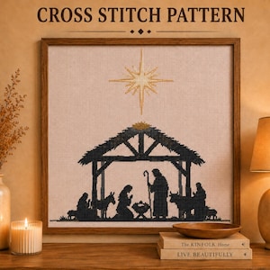 May include: A framed cross stitch pattern featuring a black silhouette of a nativity scene under a large gold star. The design includes figures of people, a donkey, and a cow inside a stable structure, all stitched on light beige fabric.