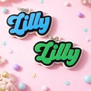 May include: Two name tags with the name "Lilly" in a playful font. One tag is blue with a black outline, the other is green with a black outline. Both have a white border and a silver keyring. The background is pink with decorative elements.