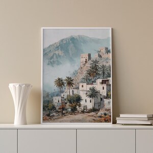 May include: Art print showing a village on a mountainside. The image features white buildings and palm trees, set against a misty mountain range. The colour palette includes white, blue, and brown hues. The print is framed in a white border.