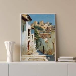May include: A framed art print showing a European street scene bathed in sunlight. The artwork features white buildings with brown roofs and a distant castle under a blue sky. The print is displayed on a white shelf with a white vase.