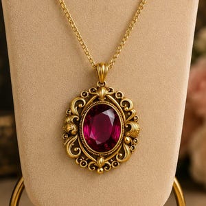 Puede incluir: Un collar de tono dorado con un colgante de piedra preciosa de color magenta intenso en forma ovalada. El colgante está engastado en un marco de metal ornamentado de estilo filigrana con detalles de volutas. El collar cuelga de una cadena fina.