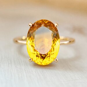 May include: A gold ring featuring a large, oval-cut yellow gemstone. The gemstone has a gradient of colors, transitioning from a deep yellow to a lighter, more transparent hue. The ring's band is a simple, polished gold.
