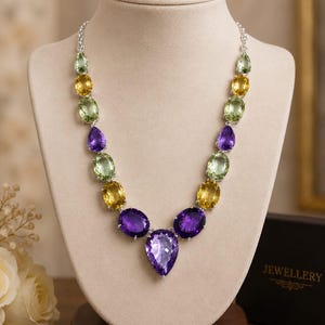 May include: A silver-toned necklace featuring a symmetrical arrangement of faceted gemstones in shades of light green, yellow, and deep purple. The stones are oval and pear-shaped, set in metal prongs, and connected by a delicate silver-toned chain.