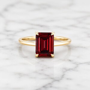 Puede incluir: Un anillo de oro con una piedra preciosa rectangular de color rojo intenso. La piedra preciosa es el punto focal, engastada en una montura de cuatro garras. La banda del anillo es de oro pulido simple. El fondo es una superficie de mármol blanco.