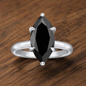 Könnte beinhalten: Ein silberner Ring mit einem großen, marquiseförmigen schwarzen Edelstein. Der facettierte Edelstein reflektiert das Licht und wird von vier Krappen gehalten. Der glatte, polierte Ring sitzt auf einer Holzoberfläche.