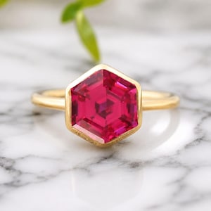 May include: A gold ring featuring a large, faceted, hexagonal ruby gemstone. The ruby is a vibrant shade of red with a complex internal structure. The ring's band is a simple, polished gold. The ring is set against a marble background.