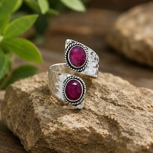 Puede incluir: Un anillo abierto de plata martillada con dos piedras preciosas de rubí de forma ovalada en un tono rojo rosado intenso. Cada piedra está engastada en un borde trenzado tipo cuerda, creando un contraste con la banda metálica texturizada.