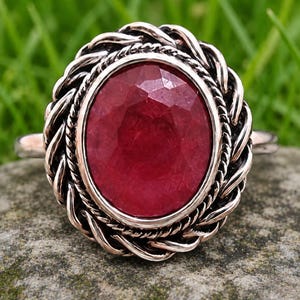 Puede incluir: Anillo de plata con una gran gema de rubí ovalada. El rubí es de color rojo intenso y está engastado en un engaste de plata trenzada decorativa. El anillo se muestra sobre una superficie de piedra sobre un fondo verde borroso.