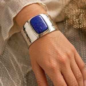Puede incluir: Un brazalete ancho de plata con textura martillada. El brazalete presenta una gran piedra preciosa rectangular de color azul oscuro en el centro. El brazalete se lleva en la muñeca, mostrando el diseño y el color de la joya.