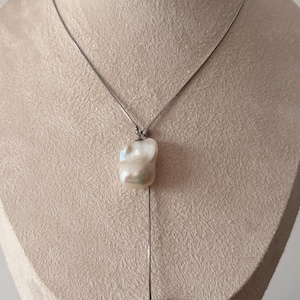 Op de afbeelding: Een zilveren ketting met een grote, onregelmatig gevormde parel hanger. De hanger is wit met iriserende tinten. Een zilveren ketting loopt van de parel en eindigt in een kleine, ronde zilveren kraal. De ketting wordt getoond op een beige buste.