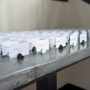 Può includere: Una collezione di segnaposto bianchi con nomi e numeri di tavolo, disposti su una superficie grigia. Ogni biglietto è tenuto da un supporto a forma di cuore, alternando bianco e nero. Per un matrimonio o evento.