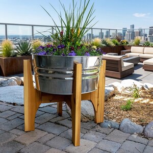 May include: A galvanized steel tub planter with horizontal ridges and side handles, supported by a four-legged wooden stand. The tub is filled with a variety of green foliage, tall grasses, and small purple and yellow flowers.