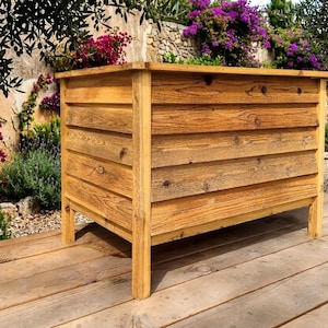 May include: A rectangular wooden planter box with a natural finish. The box is constructed from horizontal planks and has four legs. The background features a garden with flowers and greenery.