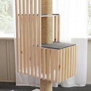 May include: A multi-level cat tree with a natural wood finish. The structure features multiple platforms, scratching posts wrapped in sisal rope, and a top perch. The design includes vertical wooden slats for a modern aesthetic. The base is a white square.