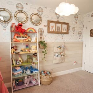 May include: A white five-tier bookshelf filled with various toys, including a red plush character, a piano toy, and stuffed animals. The room features hot air balloon wallpaper, three round mirrors, a cloud light, and a sign reading Adventure Awaits.