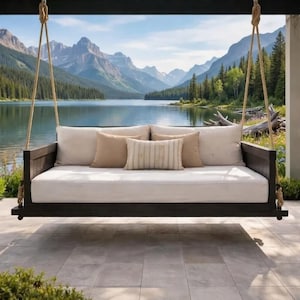 May include: A dark wood outdoor swing bed with a white cushion and several beige pillows. The swing is suspended by thick ropes. The background features a lake and mountains.