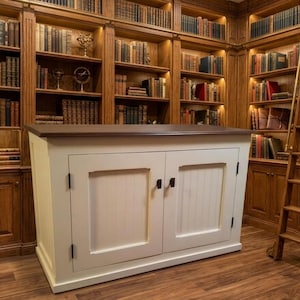 May include: A cream cabinet with a dark brown top and black handles is positioned in a library. The cabinet has two doors with vertical panels. The library has built-in bookshelves filled with books and a wooden rolling ladder.