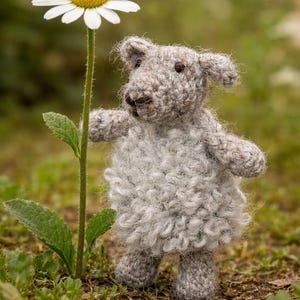 Puede incluir: Un cordero de ganchillo gris hecho a mano está de pie junto a una gran margarita con pétalos blancos y centro amarillo. El cordero tiene un cuerpo esponjoso y orejas pequeñas. La escena está ambientada al aire libre con hierba verde.