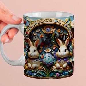 May include: White ceramic mug featuring two bunnies and a decorated Easter egg. The mug has a light blue background with floral and decorative elements. The handle is white.