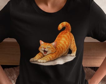 Könnte beinhalten: Schwarzes T-Shirt mit einem Cartoon-Bild einer sich streckenden, orangefarbenen Tabby-Katze mit glücklichem Gesichtsausdruck. Die Katze liegt auf einer weißen Oberfläche. Das T-Shirt hat einen Rundhalsausschnitt.
