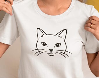 Könnte beinhalten: Weißes T-Shirt mit einem minimalistischen schwarzen Katzengesicht-Design. Die Grafik zeigt die Augen, Nase, Schnurrhaare und Ohren der Katze. Das Shirt besteht aus einem weichen Material und hat einen Rundhalsausschnitt.