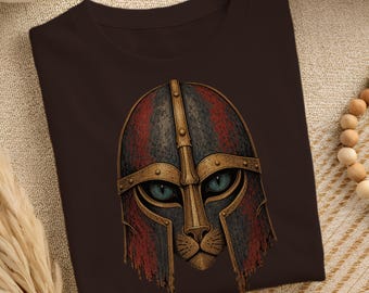 Könnte beinhalten: Ein dunkelbraunes T-Shirt mit einer grafischen Illustration eines Katzengesichts, das einen verwitterten, metallisch wirkenden Helm trägt. Der Helm hat goldfarbene Akzente und rot-blaue Texturfelder, während die Katze stechend blaue Augen hat.
