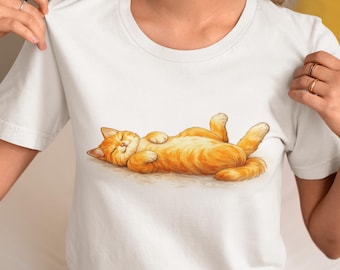 Könnte beinhalten: Weißes T-Shirt mit einer detaillierten Illustration einer entspannten, orangefarbenen Tabby-Katze, die auf dem Rücken liegt. Das Fell der Katze ist warmorange mit helleren Highlights. Das Shirt hat einen klassischen Rundhalsausschnitt.