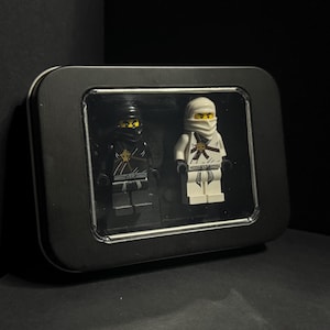 May include: A black metal display case with a clear window, containing two Lego Ninjago figures. One figure is black, the other is white. Both figures have ninja masks and gold accents. The case is on a dark surface.