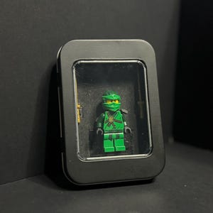 Puede incluir: Una minifigura de ninja verde con máscara y emblema dorado en el pecho, exhibida dentro de una lata de metal negra rectangular con una ventana de plástico transparente. La figura está sobre una base negra, con una espada dorada a la izquierda.
