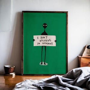 Op de afbeelding: Een ingelijste kunstafdruk met een groene achtergrond, met een zwart alienfiguur dat een bord vasthoudt. Op het bord staat "I DON'T BELIEVE IN HUMANS" in zwarte letters. Het kunstwerk wordt getoond in een kamer met een houten vloer en een bed.