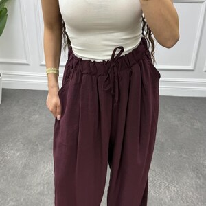 Op de afbeelding: Een mouwloze witte top gecombineerd met een wijde, bordeauxrode broek. De broek heeft een taille met trekkoord en zakken. De outfit is geaccentueerd met gouden armbanden. Het model draagt sandalen.