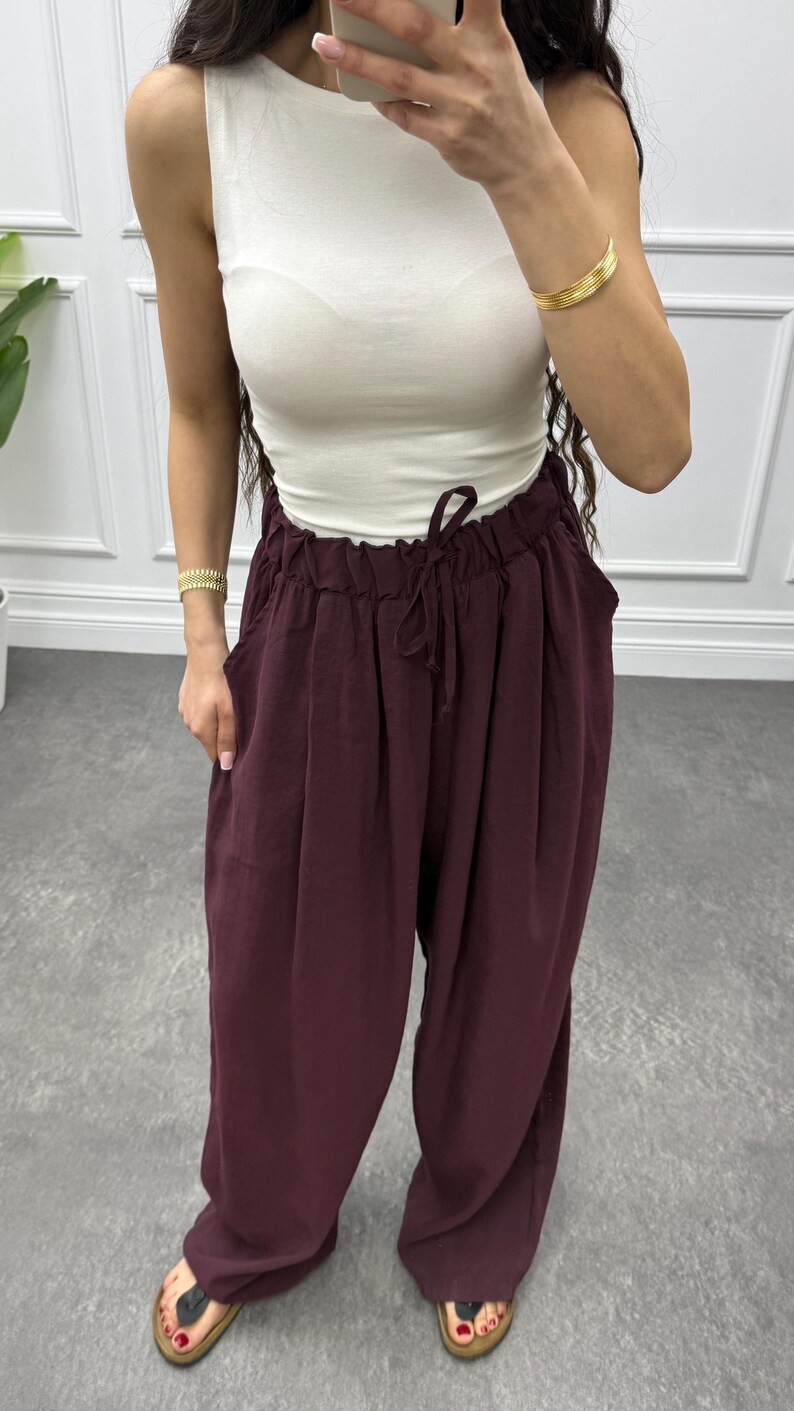 Op de afbeelding: Een mouwloze witte top gecombineerd met een wijde, bordeauxrode broek. De broek heeft een taille met trekkoord en zakken. De outfit is geaccentueerd met gouden armbanden. Het model draagt sandalen.