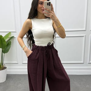 Op de afbeelding: Een vrouw draagt een witte mouwloze top en een wijde, bordeauxrode broek. De broek heeft een taille met trekkoord en zakken. Ze houdt een telefoon vast en draagt gouden armbanden.